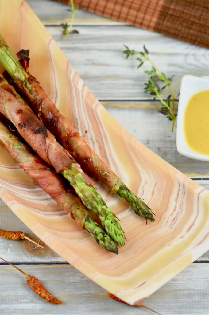 Grüner Spargel in Schinken gegrillt auf länglicher Schale aus Onyxmarmor. Hintergrund hell. Hochkant