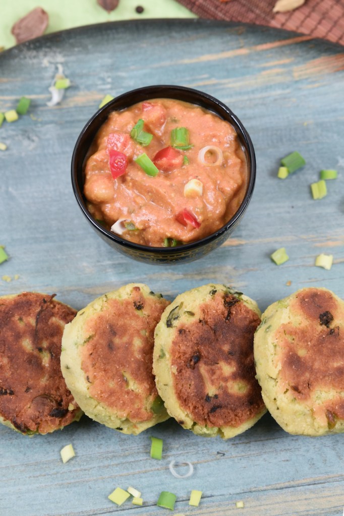 Indische Kartoffelküchlein auf blauem Teller. Dahinter steht eine Schale mit Kichererbsen-Curry.