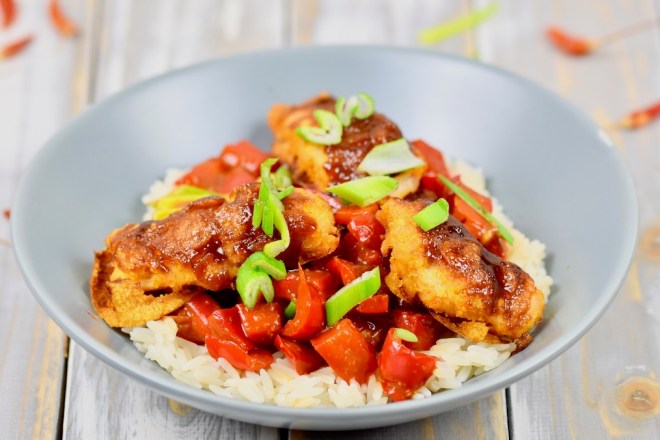 Gebackenes Hoisin-Hähnchen auf Paprikagemüse mit Reis in hellgrauem Teller. Der Teller steht auf einer weißen Holzfläche.