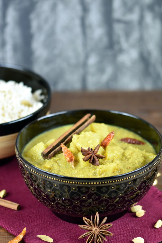Veganes Kartoffelcurry in einer schwarzen Schale. Im Hintergrund steht der Kokosreis in einer anderen asiatischen Schale.