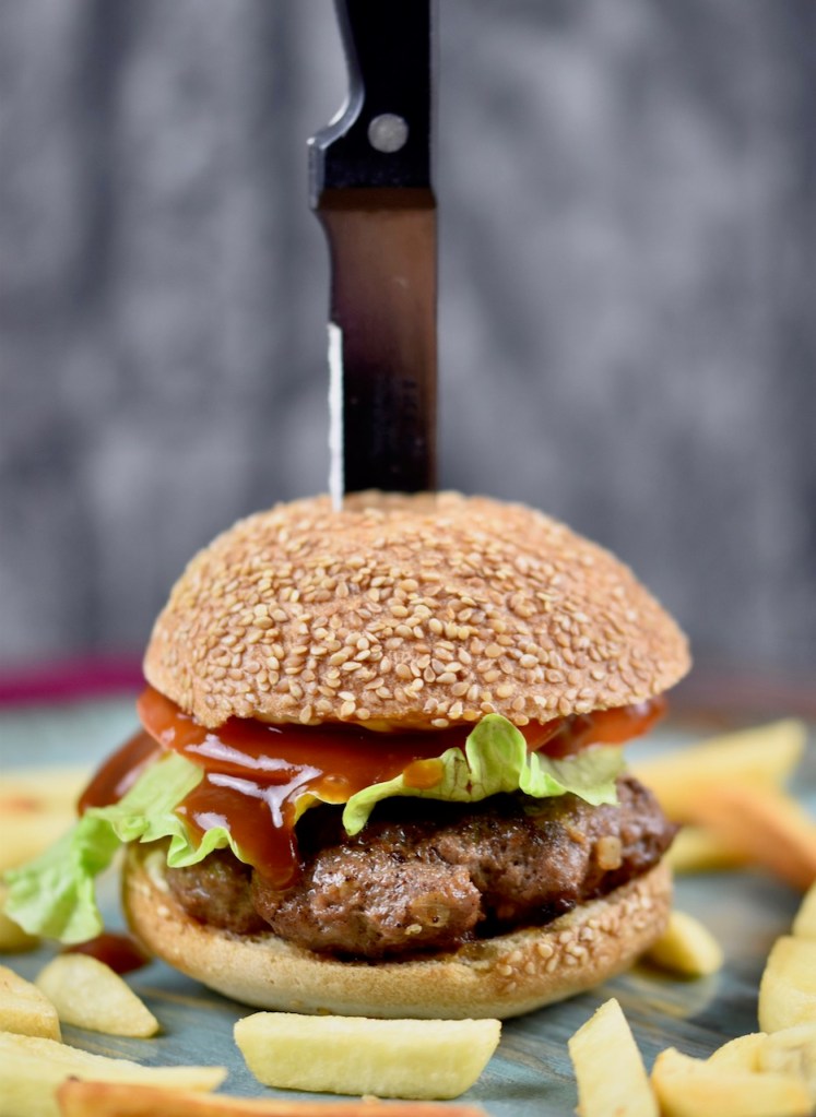 Pljescavica Burger aufgespießt mit einem Messer auf einem bläulichen Teller. Drumherum liegen Pommes frites. Dunkler Hintergrund.