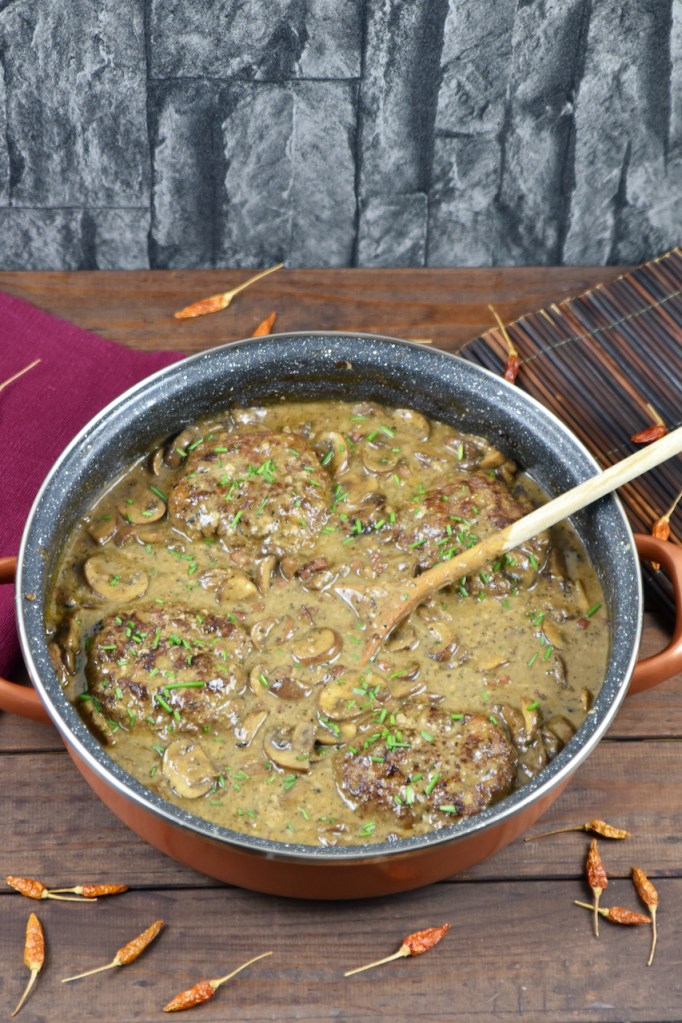 Frikadellen aus dem Backofen in cremiger Pilzsauce in einer tiefen Pfanne. Hintergrund dunkel.