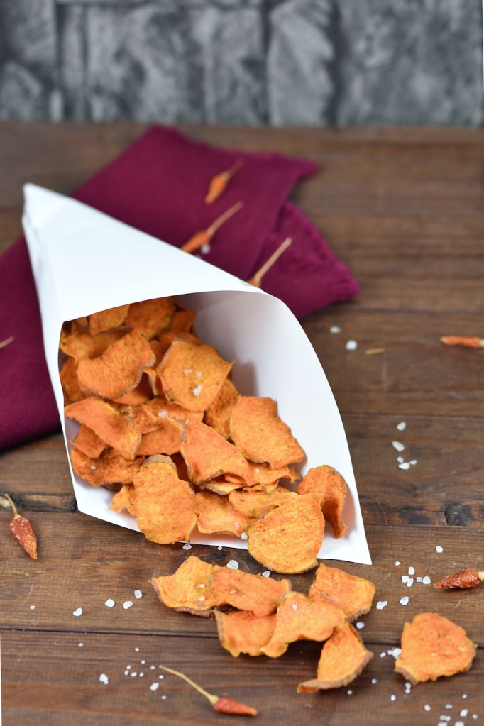 Knusprige Süßkartoffelchips mit BBQ-Aroma in einer Papiertüte. Hintergrund dunkel.