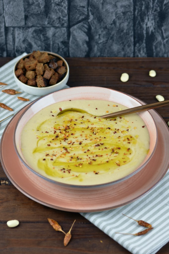 Cremige Blumenkohlsuppe in einem rosa Teller. Im Hintergrund stehen die Croûtons.