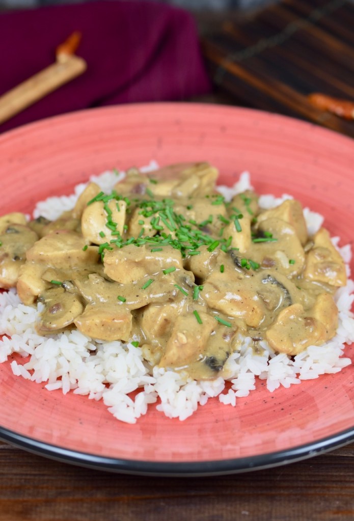 Hähnchen-Geschnetzeltes mit Pilzen auf Reis auf einem roten Teller, dekoriert mit Schnittlauchröllchen. Hintergrund dunkel