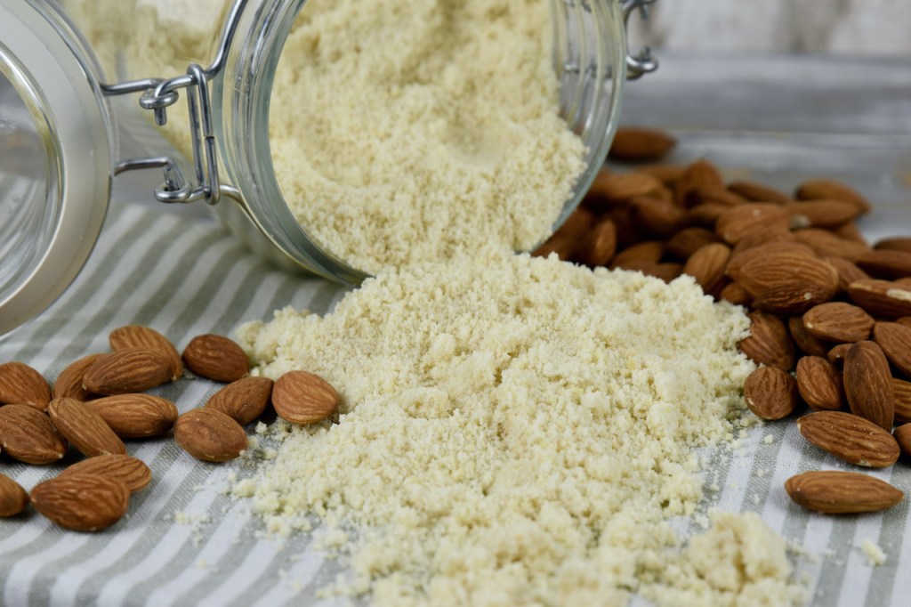 Mandelmehl aus einem liegenden Vorratsglas teilweise ausgekippt. daneben liegen ganze Mandeln ohne harte Schale.