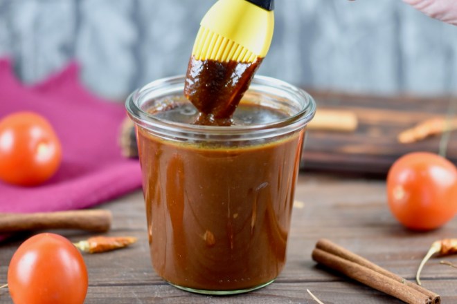Rauchige BBQ-Sauce in einem Glas. Ein Pinsel mit gelben Silikonborsten wird in die Sauce getunkt. Hintergrund dunkel.