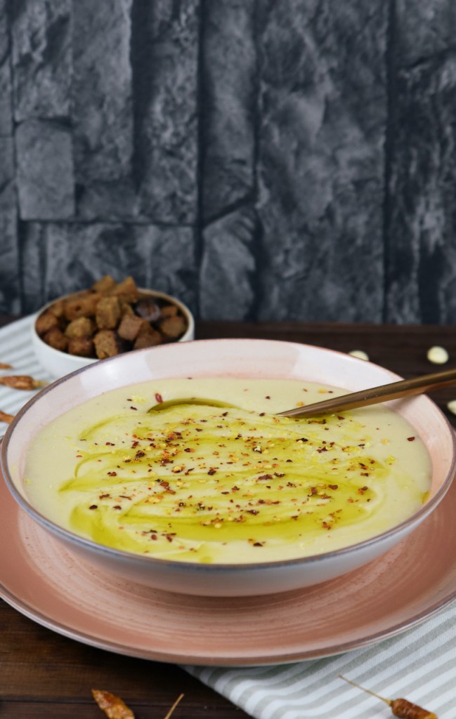 Cremige Blumenkohlsuppe in einem rosa Teller. Im Hintergrund stehen die Croûtons.