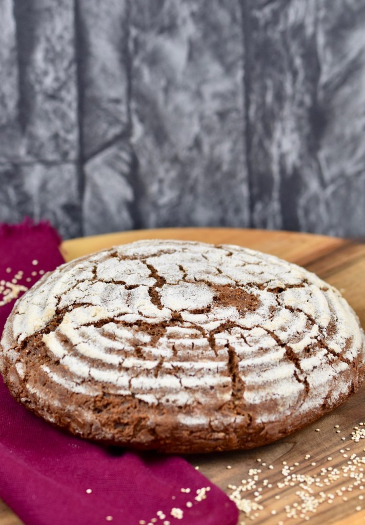 glutenfreies Krustenbrot in rund auf Holzbrett. Dekoriert ist es mit einer weinroten Stoffserviette und groben Salzkörnern.