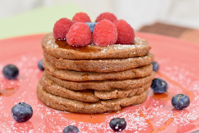 Glutenfreie Pfannkuchen gestapelt auf roten Teller, garniert mit Himbeeren, Blaubeeren, Ahornsirup und Puderzucker. Hintergrund hell.