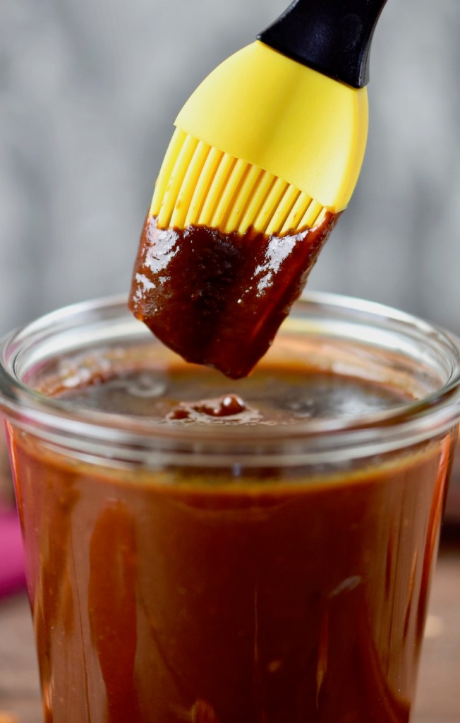 Rauchige BBQ-Sauce in einem Glas. Ein Pinsel mit gelben Silikonborsten wird in die Sauce getunkt. Hintergrund dunkel.