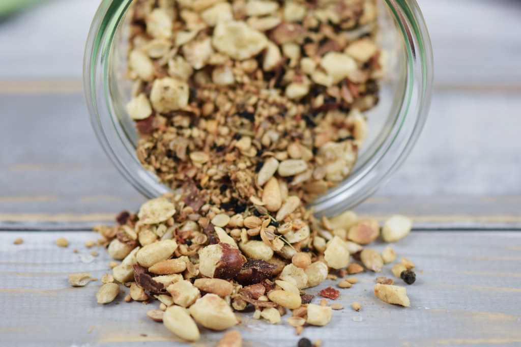 Dukkah selber gemacht in einem liegenden Glas. Etwas Dukkah ist herausgerutscht. Hintergrund hell.