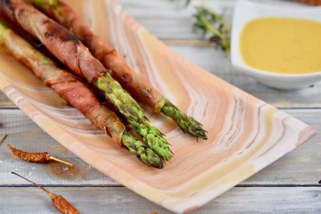 Grüner Spargel in Schinken gegrillt auf länglicher Schale aus Onyxmarmor. Hintergrund hell.