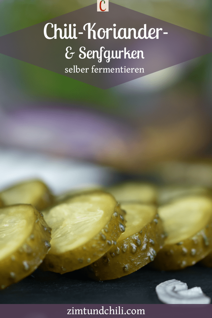 Im Hintergrund sind fermentierte Gurken in einem Einmachglas. Davor liegen die Senfgurken angeschnitten auf einer Schieferplatte. Beschrifteter Pin.