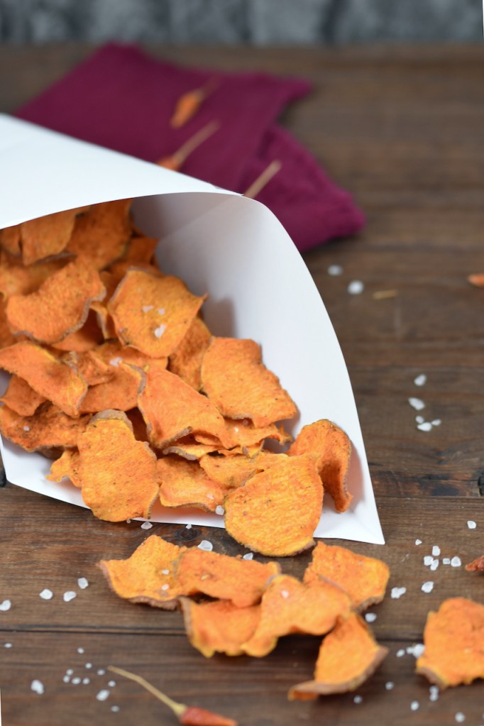 Knusprige Süßkartoffelchips mit BBQ-Aroma in einer Papiertüte. Hintergrund dunkel.