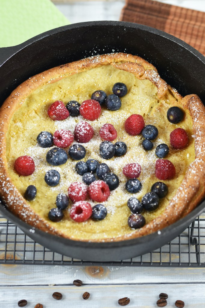 Glutenfreier Dutch Baby im schwarzen Gusseisentopf, dekoriert mit Himbeeren, Blaubeeren und Puderzucker. Hintergrund hell.
