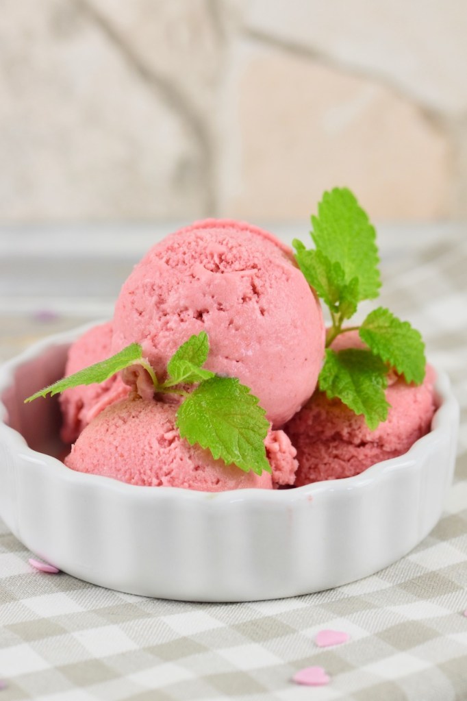 Veganes Erdbeereis selber machen - 4 Kugeln Erdbeereis in einer weißen Schale garniert mit Zitronenmelisse. Hintergrund hell.