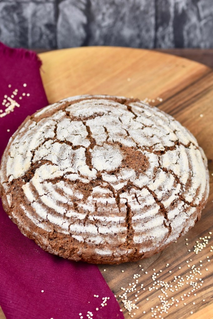 glutenfreies Krustenbrot in rund auf Holzbrett. Dekoriert ist es mit einer weinroten Stoffserviette und groben Salzkörnern.
