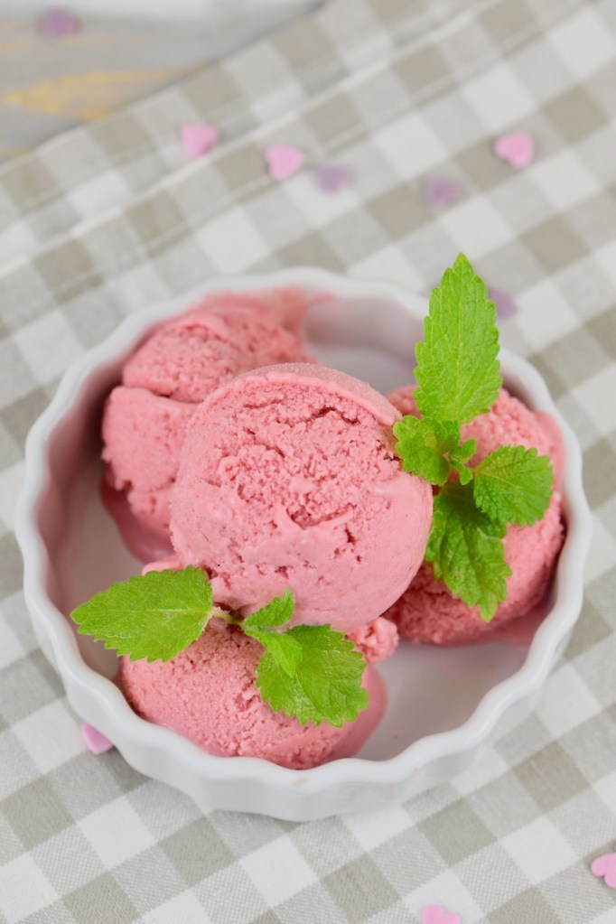 Veganes Erdbeereis selber machen - 4 Kugeln Erdbeereis in einer weißen Schale garniert mit Zitronenmelisse. Hintergrund hell.