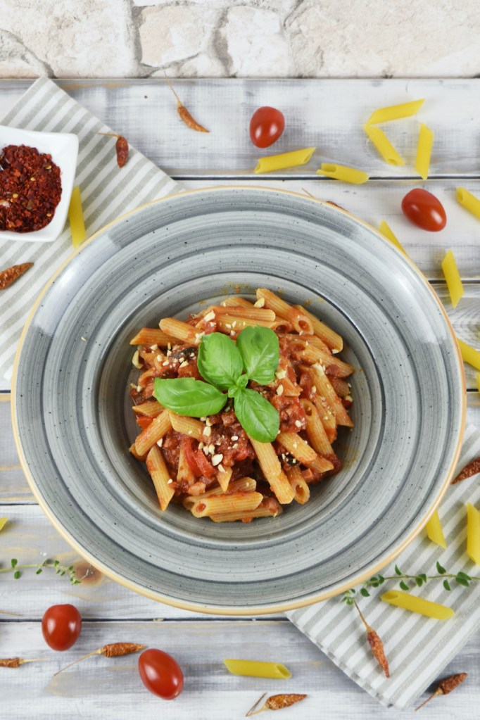 Penne Arrabiata mit Paprika und Oliven auf einem grauen Teller. Hintergrund hell.