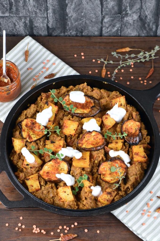 Linsen mit Harissa Kartoffeln und Auberginen mit ein paar Klecksen Joghurt dekoriert in einer schwarzen Pfanne aus Gusseisen.