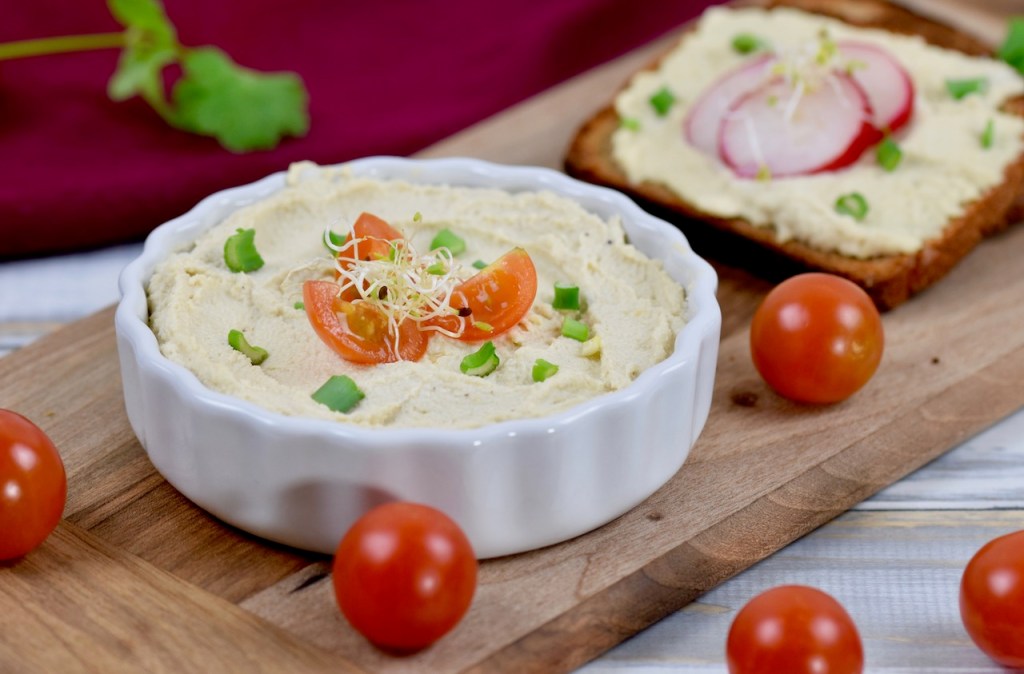 Veganer Frischkäse in weißer Schale, die auf einem Holzbrettchen steht. Der Frischkäse ist mit Vierteln von Cocktailtomaten, Frühlingszwiebel und Sprossen garniert. Im Hintergrund liegt eine Scheibe Brot mit Frischkäse und drei Scheiben Radieschen.