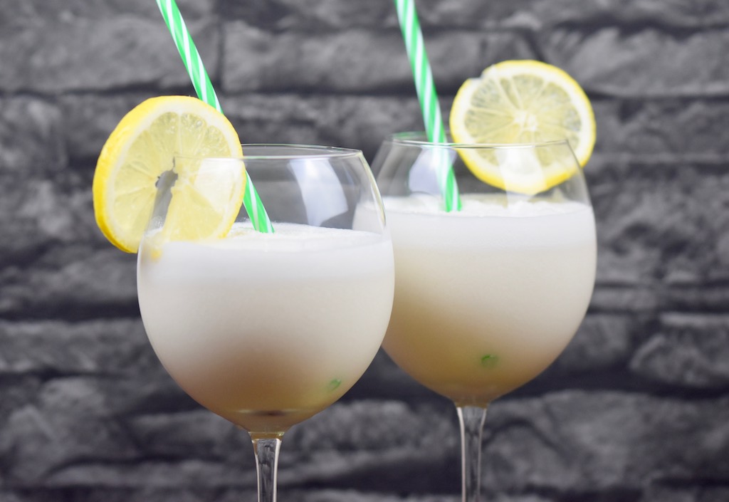 Tropische Kokoslimonade in zwei Weingläsern garniert mit einer Zitronenscheibe und einem Strohhalm. Hintergrund dunkel.