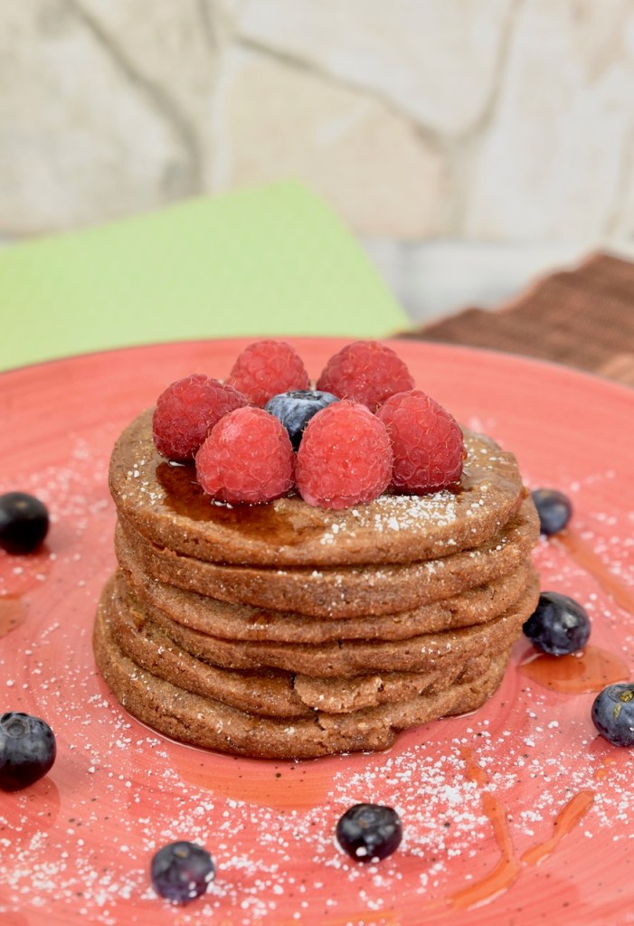 Glutenfreie Pfannkuchen gestapelt auf roten Teller, garniert mit Himbeeren, Blaubeeren, Ahornsirup und Puderzucker. Hintergrund hell.