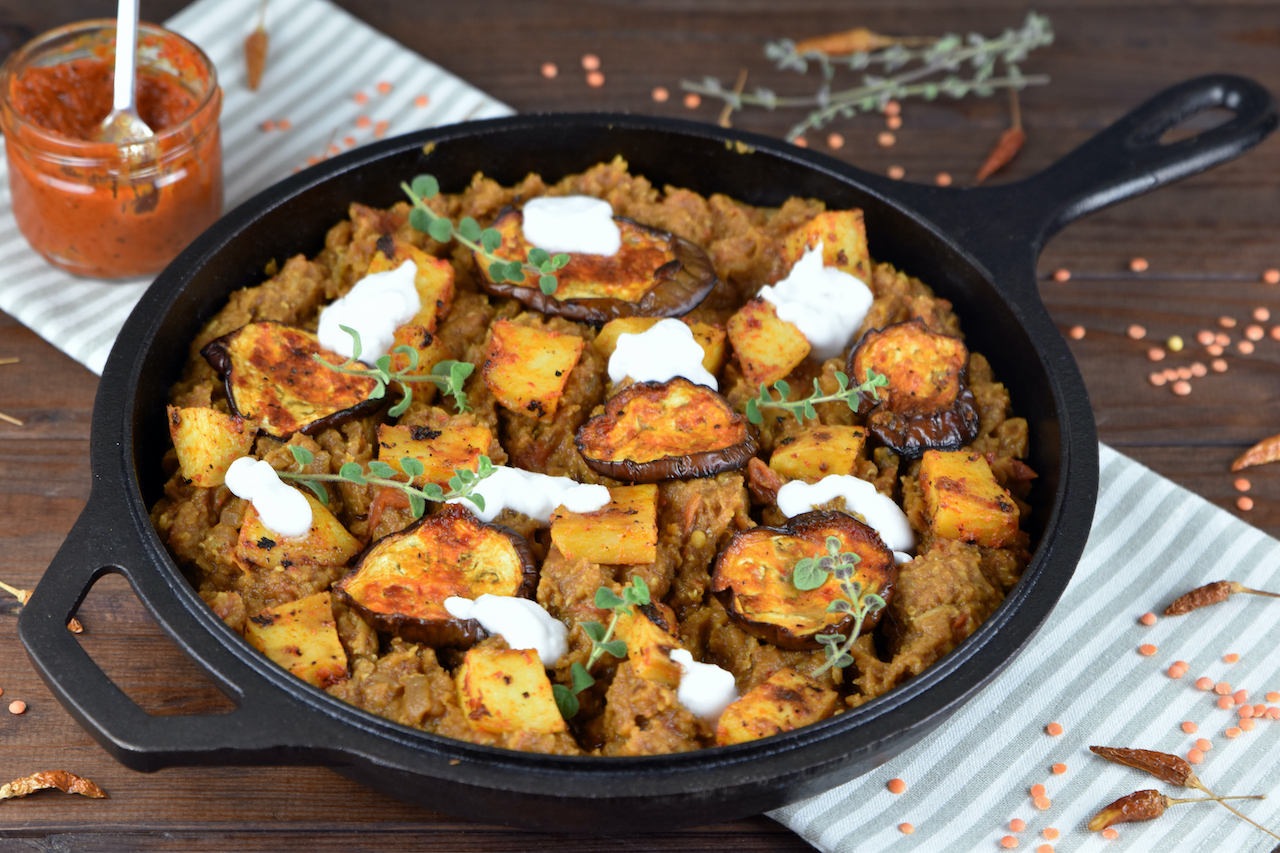 Linsen mit Harissa Kartoffeln und Auberginen mit ein paar Klecksen Joghurt dekoriert in einer schwarzen Pfanne aus Gusseisen.