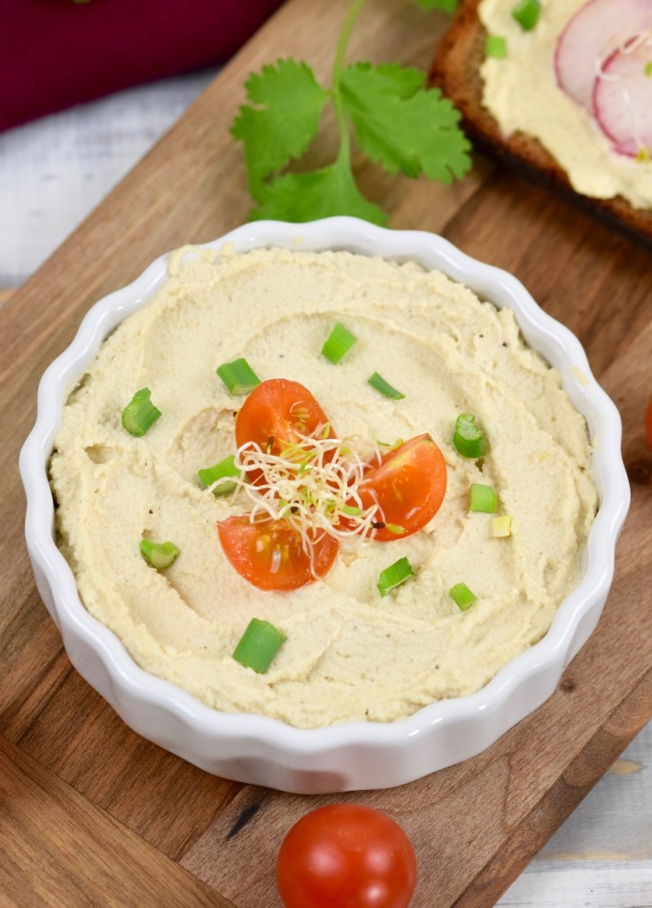 Veganer Frischkäse in weißer Schale, die auf einem Holzbrettchen steht. Der Frischkäse ist mit Vierteln von Cocktailtomaten, Frühlingszwiebel und Sprossen garniert. Im Hintergrund liegt eine Scheibe Brot mit Frischkäse und Radieschen. Nahaufnahme