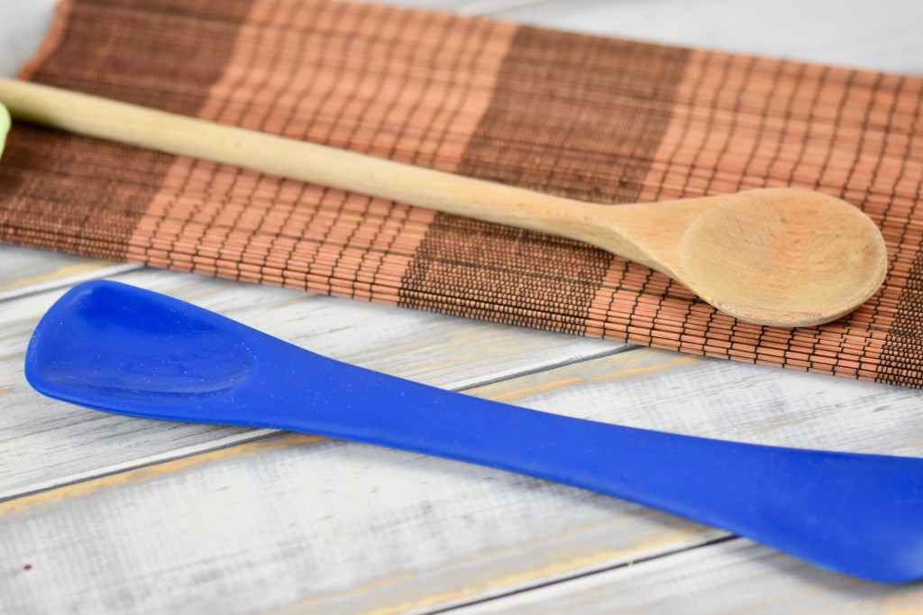 Silikonschaber und Holzlöffel liegen auf einem weißen Holztisch, Teilweise sieht man eine braunes Tischset.