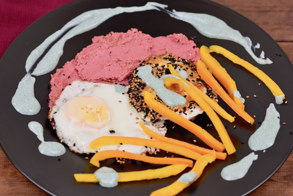 Knusprige Spiegeleier mit einer Kruste aus Sesam und Schwarzkümmelsamen mit pinkem Rote Beete Hummus und grüner Tahini-Sauce. Dazu gibt es gelbe Paprikastreifen Angerichtet ist alles auf einem schwarzen Teller.