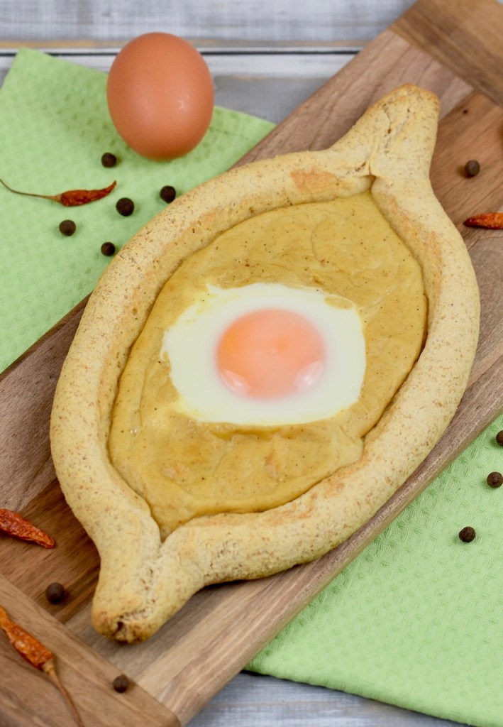 Chatschapuri mit Ei und Senfsauce auf länglichem Holzbrettchen. Heller Hintergrund dekoriert mit einem Ei, getrockneten Chilischoten und Pimentkörnern.