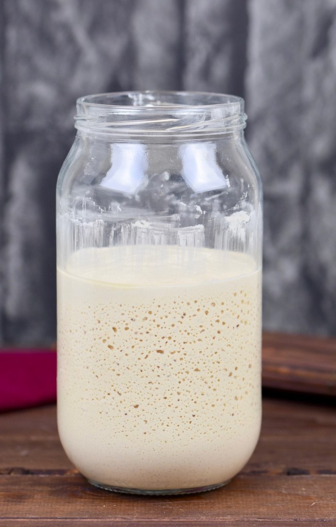 Glutenfreier Sauerteig im Glas. Man sieht die Bläschenbildung. Hintergrund dunkel.
