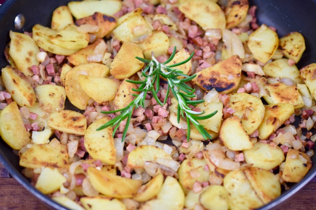 Knusprige Bratkartoffeln mit Speck und Zwiebeln in einer Pfanne, garniert mit Rosmarin - Nahaufnahme
