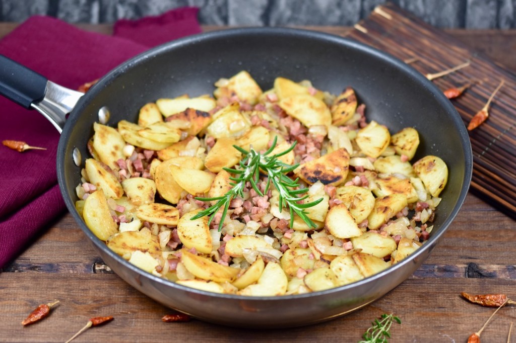 Knusprige Bratkartoffeln mit Speck und Zwiebeln in einer Pfanne, garniert mit Rosmarin