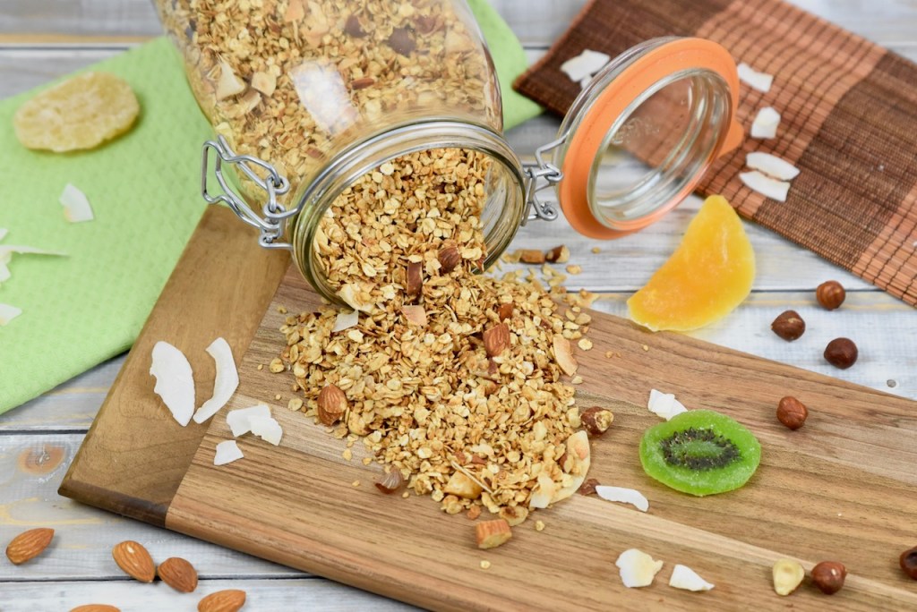 Veganer Ernährungsplan - Knuspriges Granola, das aus einem liegenden Glas geschüttet wurde. Das ausgeschüttete Granola liegt auf einem Holzbrett und der Hintergrund ist hell.