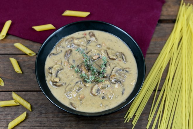 Cremige Pilzsauce aus Cashewkernen in einer schwarzen Schüssel. Drumherum liegen italienische Pasta. Hintergrund dunkel.