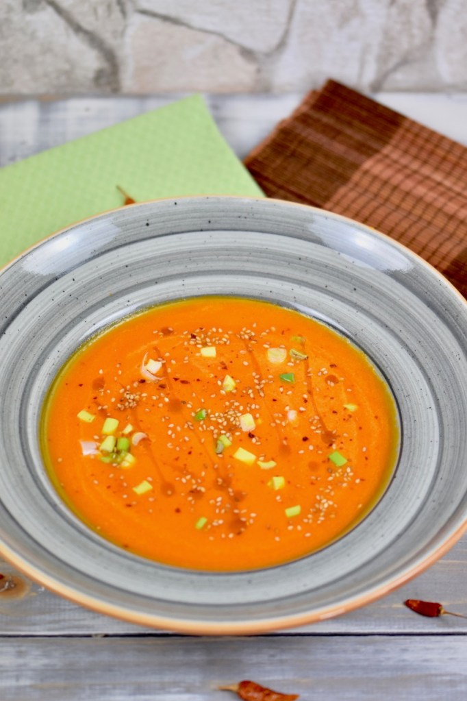 Karottensuppe mit Ingwer und Orange in grauem Teller. Garniert mit Sesamsamen, Sesamöl und Frühlingszwiebeln. Hintergrund hell.