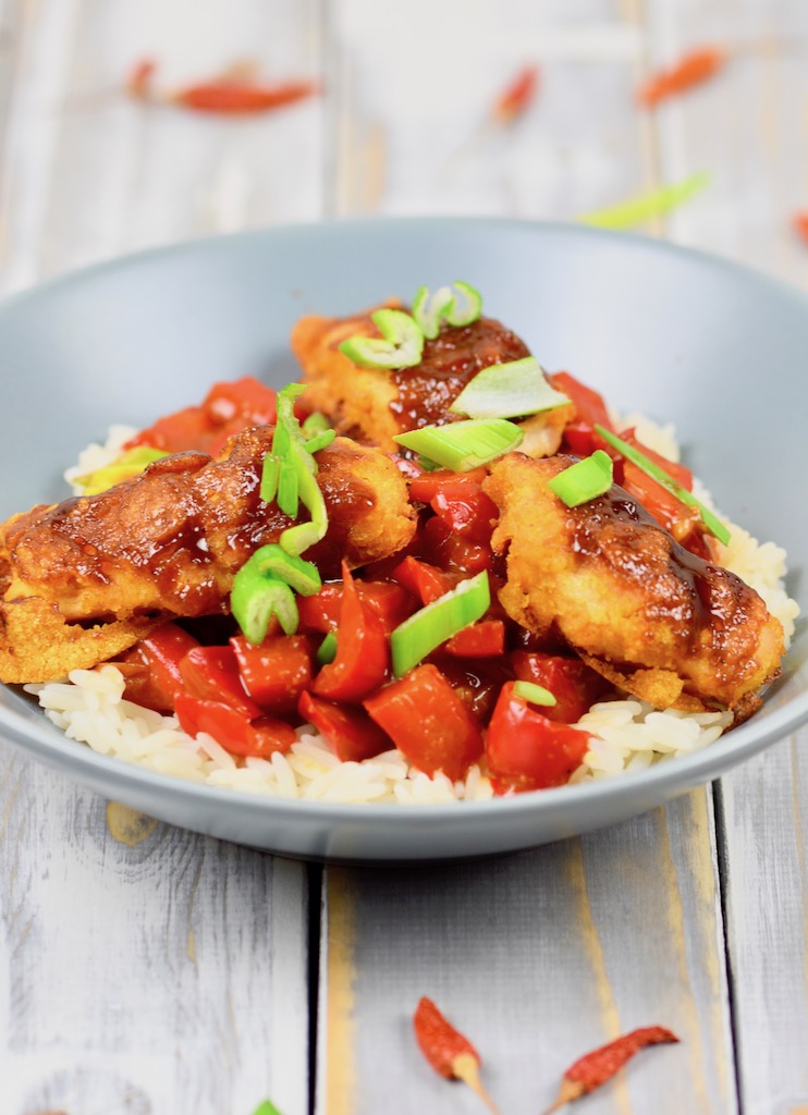 Gebackenes Hoisin-Hähnchen auf Paprikagemüse mit Reis in hellgrauem Teller. Der Teller steht auf einer weißen Holzfläche.