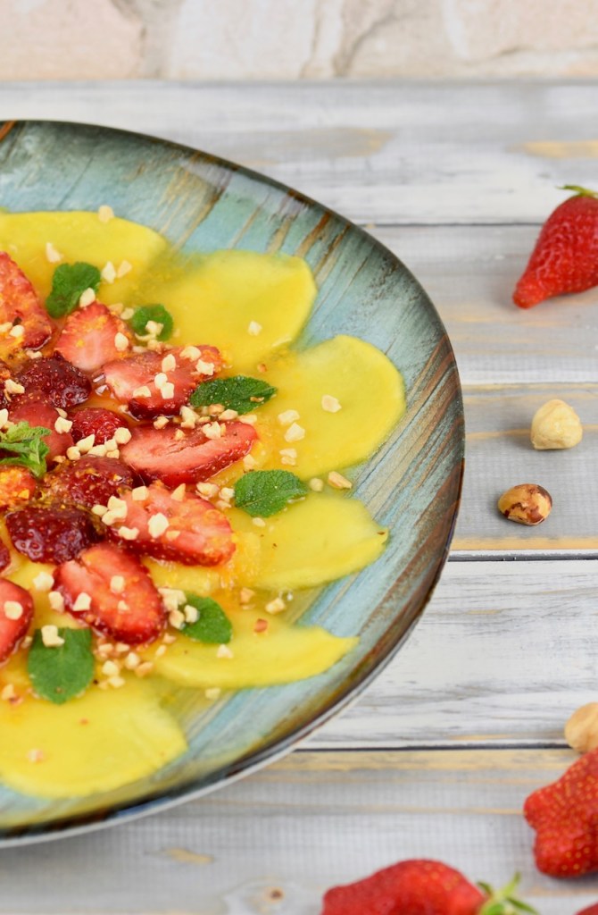 Erdbeer-Mango-Carpaccio mit Minze und Orangen-Dressing auf türkisem Teller garniert mit Minze und Haselnusskernen. Hintergrund weiß. Teller nur halb zu sehen.