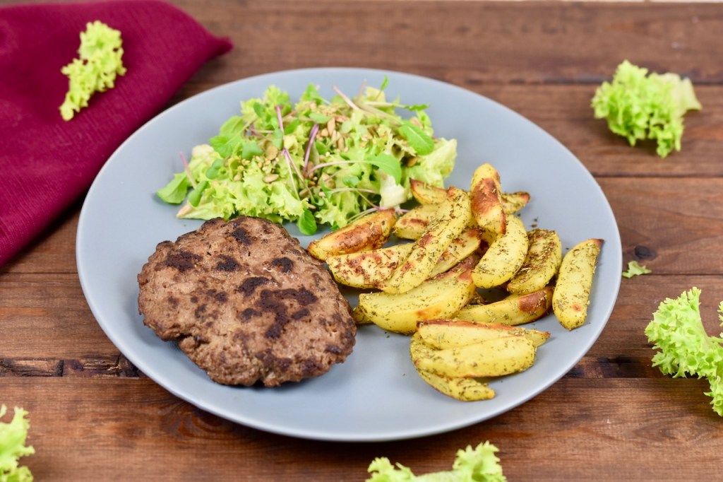 Pljescavica als Tellergericht mit Salat und Kartoffelspalten - auf grauem Teller, der auf einer dunklen Holzplatte steht