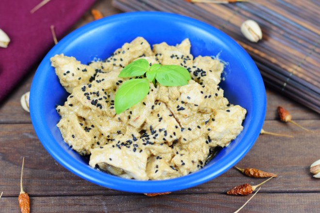 Hähnchen-Mezze in Tahini-Kokossauce in einer blauen Schale garniert mit Basilikum. Hintergrund dunkel.