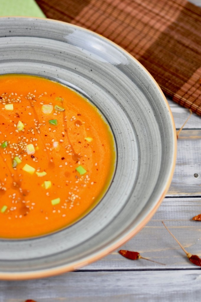 Karottensuppe mit Ingwer und Orange in grauem Teller. Garniert mit Sesamsamen, Sesamöl und Frühlingszwiebeln. Der Teller ist nur halb zu sehen. Hintergrund hell.