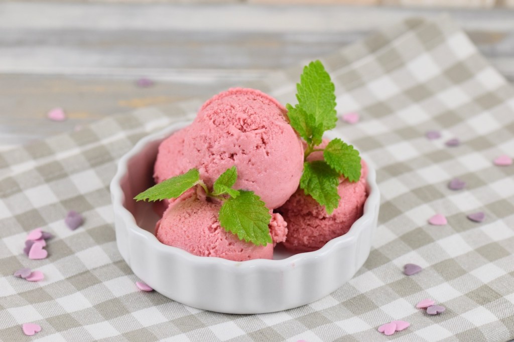 Veganes Erdbeereis selber machen - 4 Kugeln Erdbeereis in einer weißen Schale garniert mit Zitronenmelisse. Hintergrund hell.