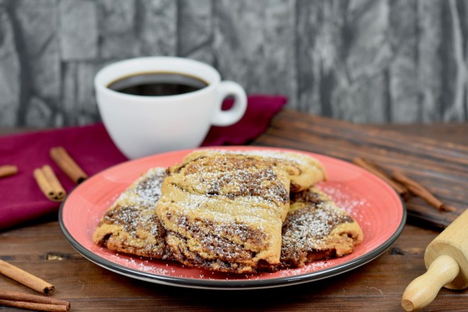 Glutenfreie Franzbrötchen auf rotem Teller. Im Hintergrund sieht man Zimtstangen, eine weiße Tasse gefüllt mit Kaffee und ein Stück von einem Nudelholz. Der Hintergrund ist dunkel.