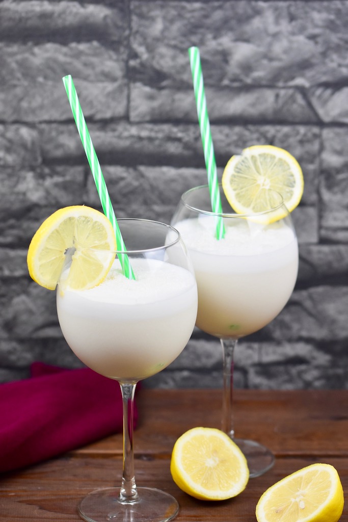 Tropische Kokoslimonade in zwei Weingläsern garniert mit einer Zitronenscheibe und einem Strohhalm. Hintergrund dunkel.