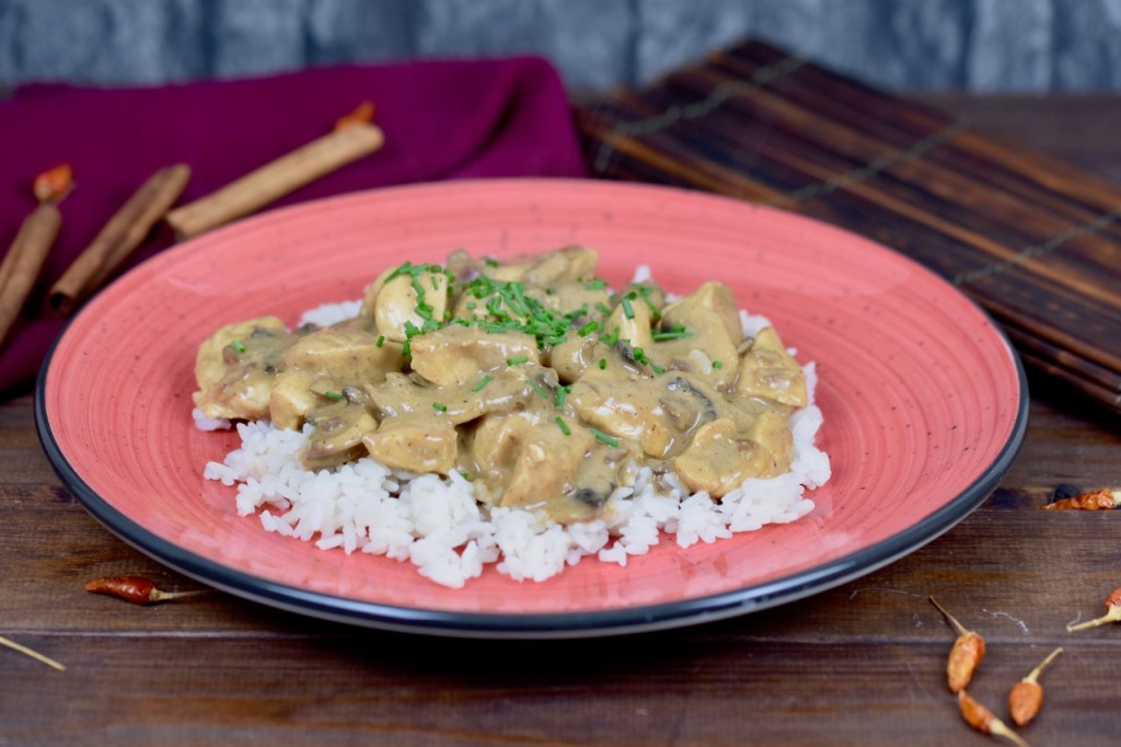 Hähnchen-Geschnetzeltes mit Pilzen auf Reis auf einem roten Teller, dekoriert mit Schnittlauchröllchen. Hintergrund dunkel