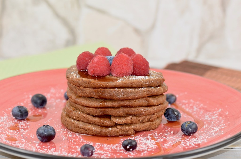 Glutenfreie Pfannkuchen gestapelt auf roten Teller, garniert mit Himbeeren, Blaubeeren, Ahornsirup und Puderzucker. Hintergrund hell.