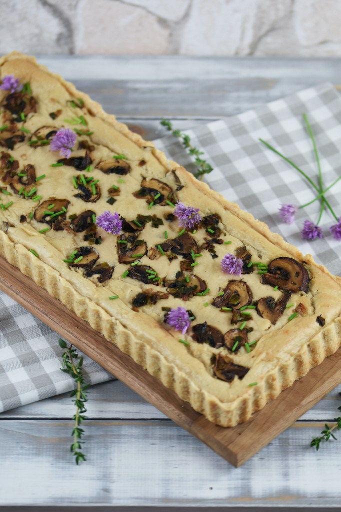 Längliche Quiche mit Lauch in Pilzen, garniert mit Schnittlauchblüten. Hintergrund hell.